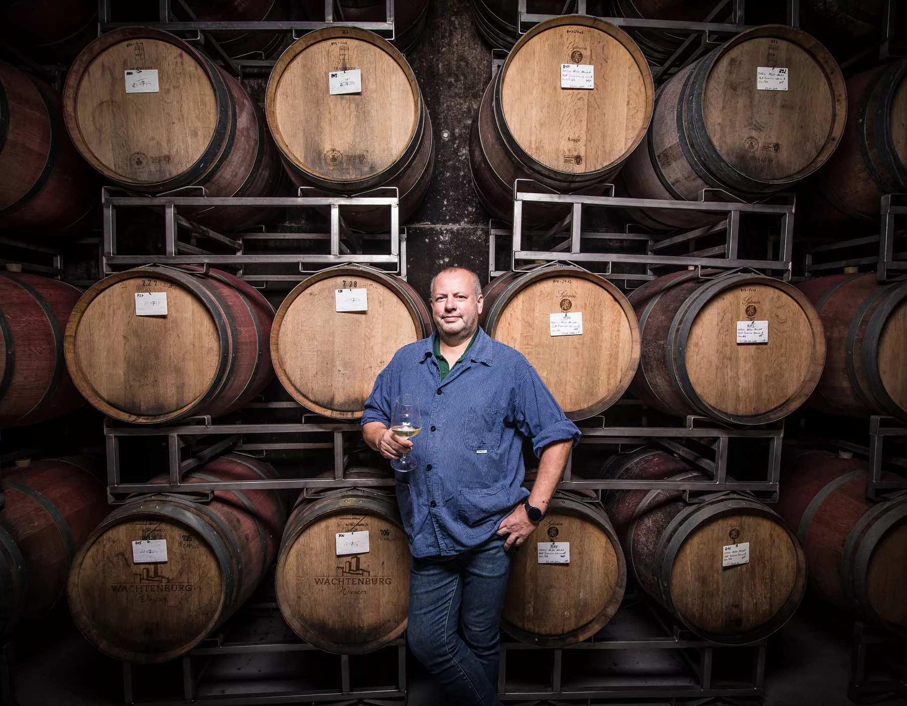 Ein Mann in einem blauen Hemd hält ein Weinglas und steht vor gestapelten hölzernen Weinfässern in einem Keller. Die Fässer sind in Reihen angeordnet und mit Schildern beschriftet.