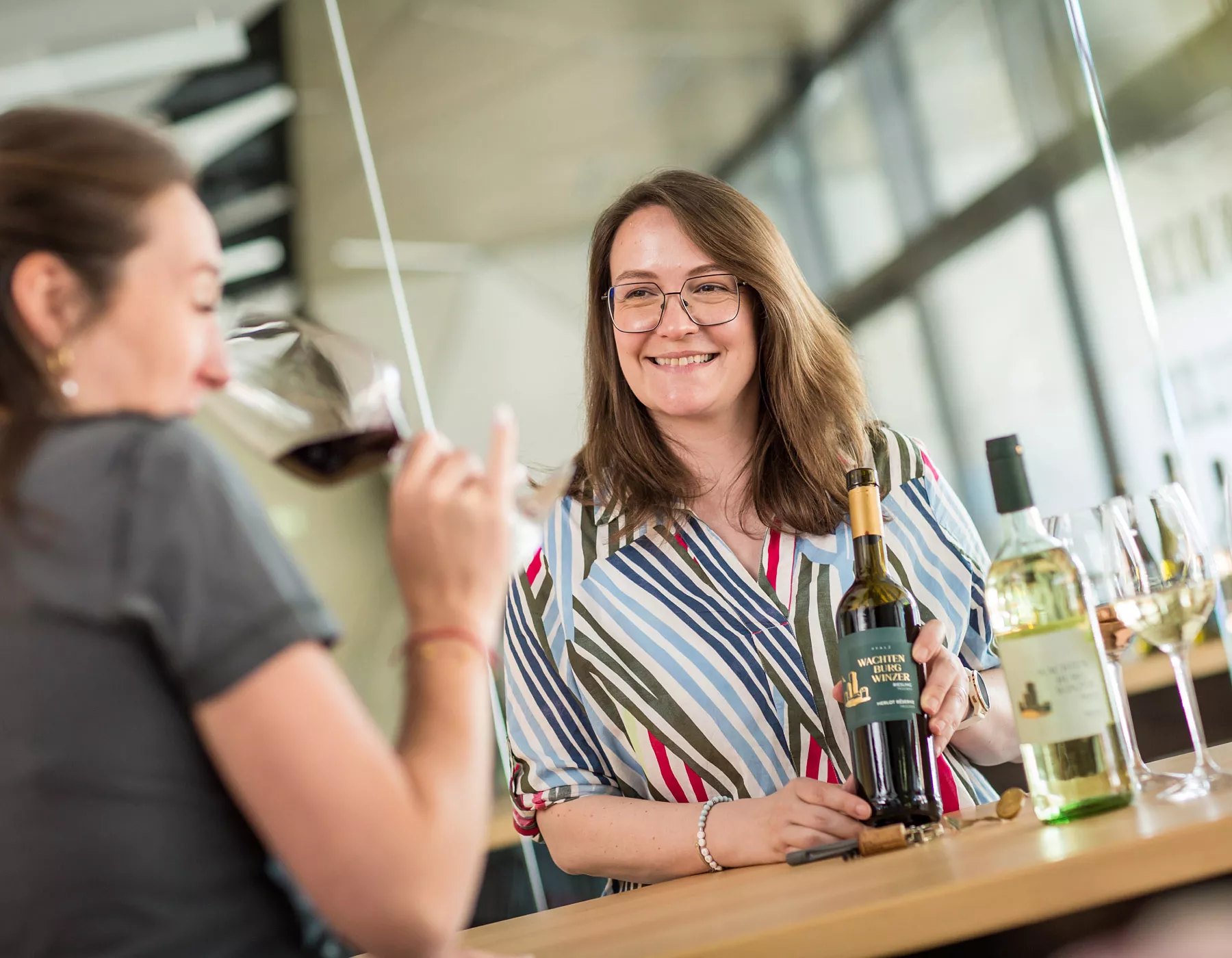 Zwei Frauen genießen eine Weinprobe; die eine trinkt aus einem Glas Rotwein, während die andere lächelt und eine Flasche Weißwein in der Hand hält. Mehrere Weingläser und Flaschen stehen auf dem Tisch vor ihnen.